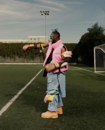Man wearing a pink puffer vest, boxing gloves, and protective pads standing on an outdoor turf sports field.