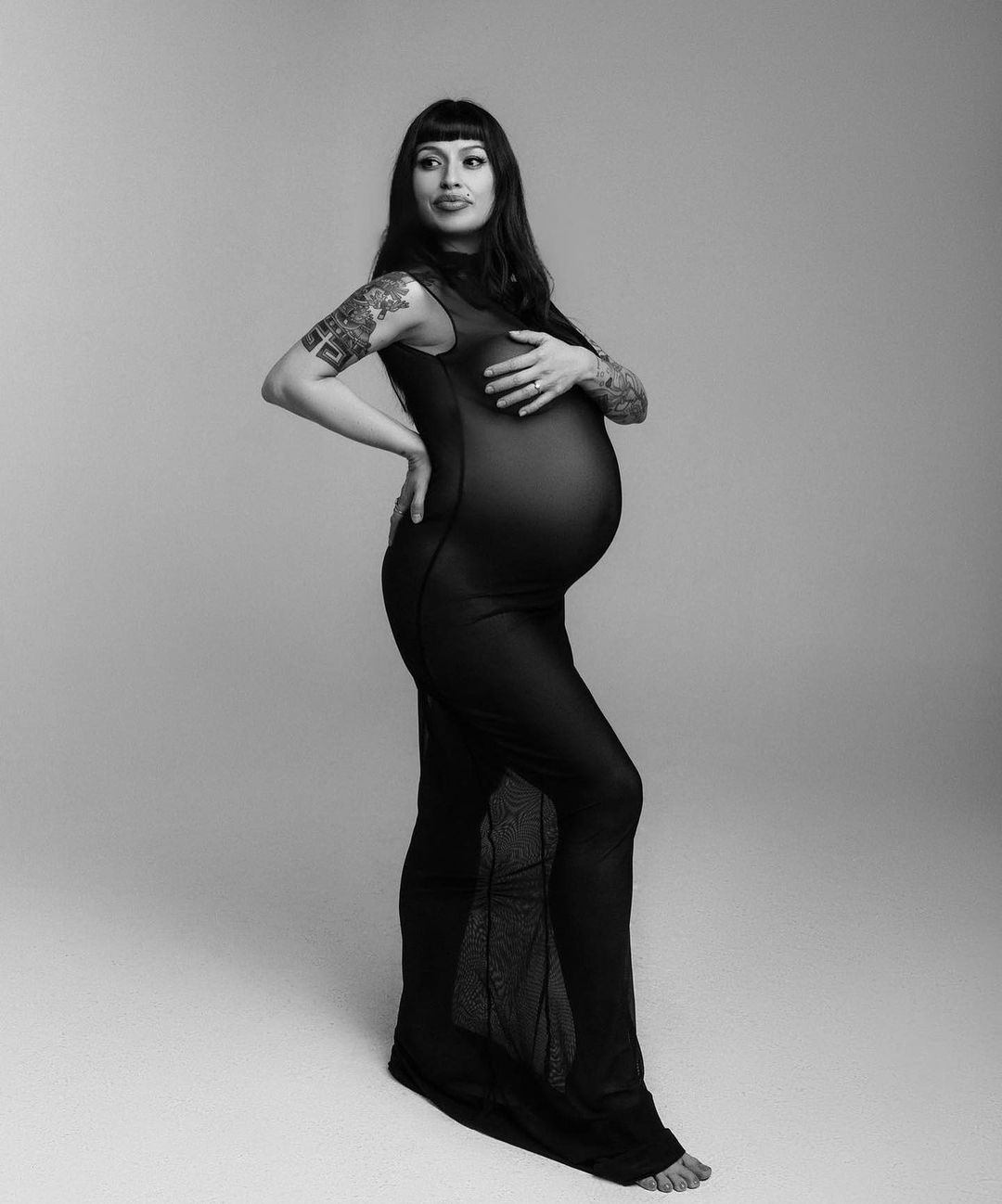 Monochrome studio portrait of a pregnant adult female with long dark hair in a sheer black sleeveless dress posing against a light background.