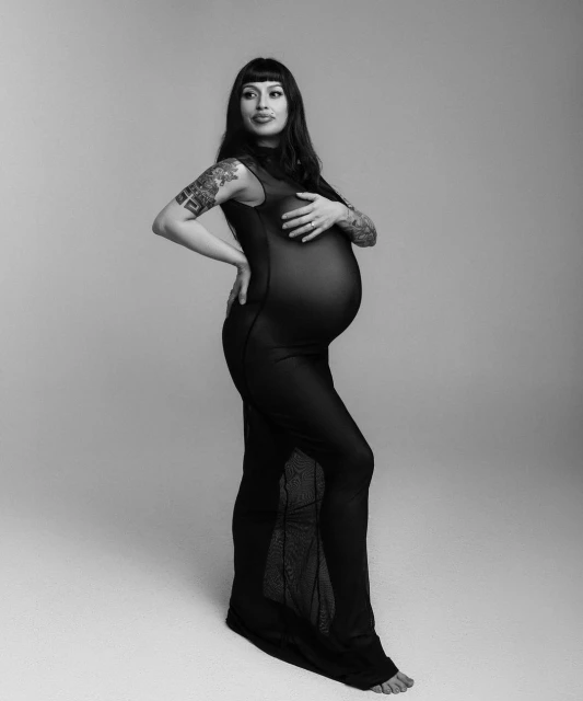 Monochrome studio portrait of a pregnant adult female with long dark hair in a sheer black sleeveless dress posing against a light background.