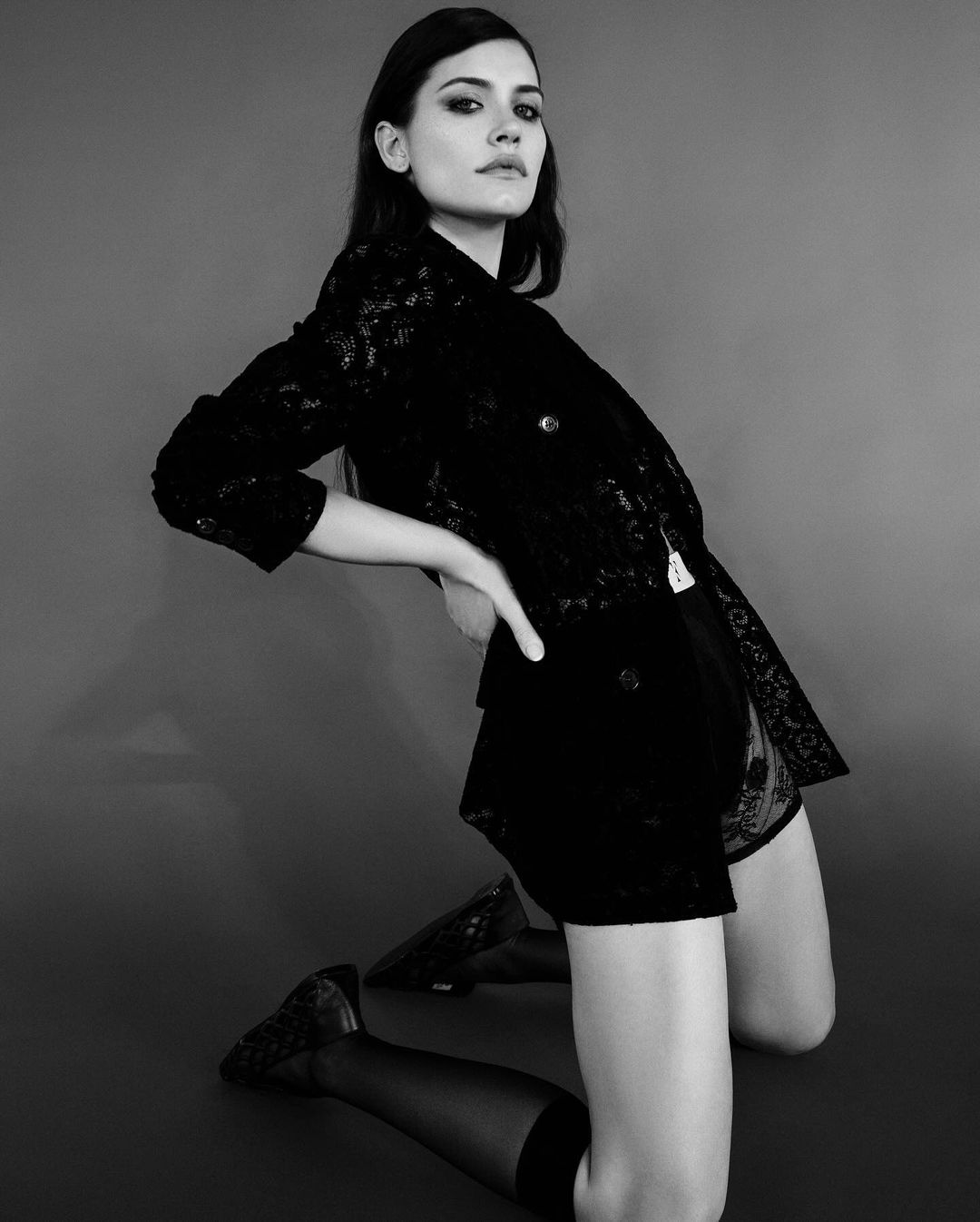 Monochrome portrait of a woman kneeling in a dark patterned blazer and shorts against a grey backdrop.