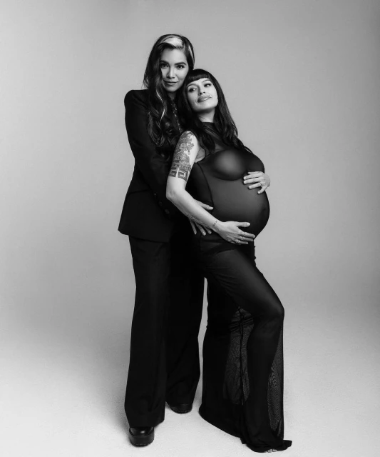 Black and white studio portrait of pregnant woman in sheer dress with partner in suit, arms around the pregnant woman.