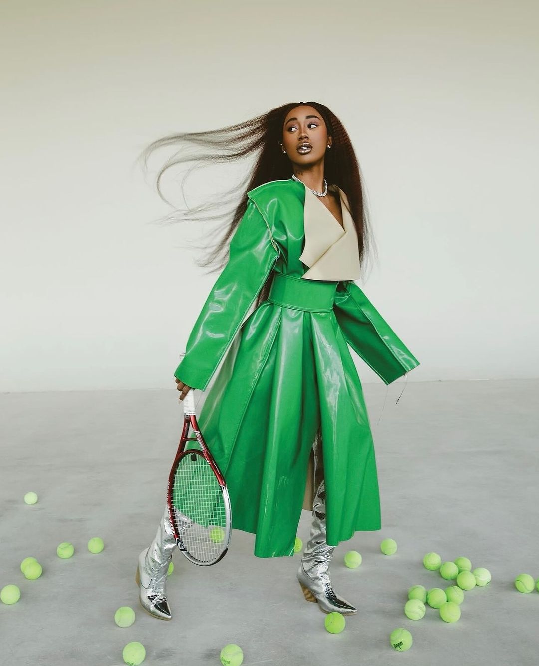 An adult woman poses indoors surrounded by tennis balls, holding a tennis racket, wearing a green coat and silver boots.