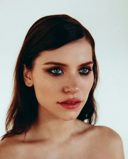 Close-up portrait of a young adult woman with dark hair and green eyes, featuring smokey eyeshadow and red lipstick, against a white background.