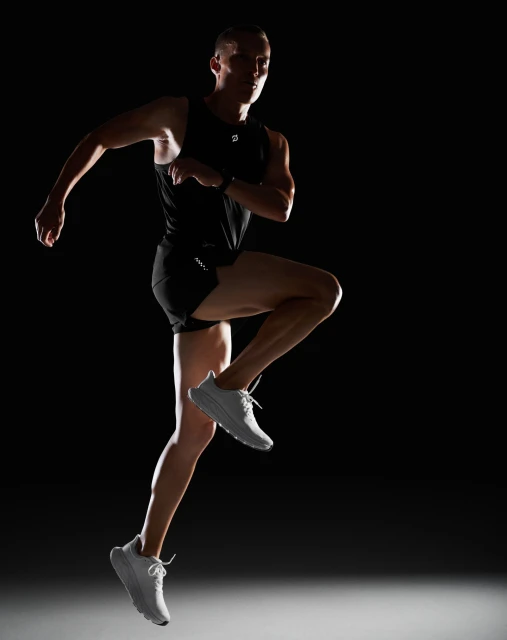Athletic man in black activewear mid-stride against a dark backdrop, showcasing fitness and athleticism, highlighted by stark lighting.