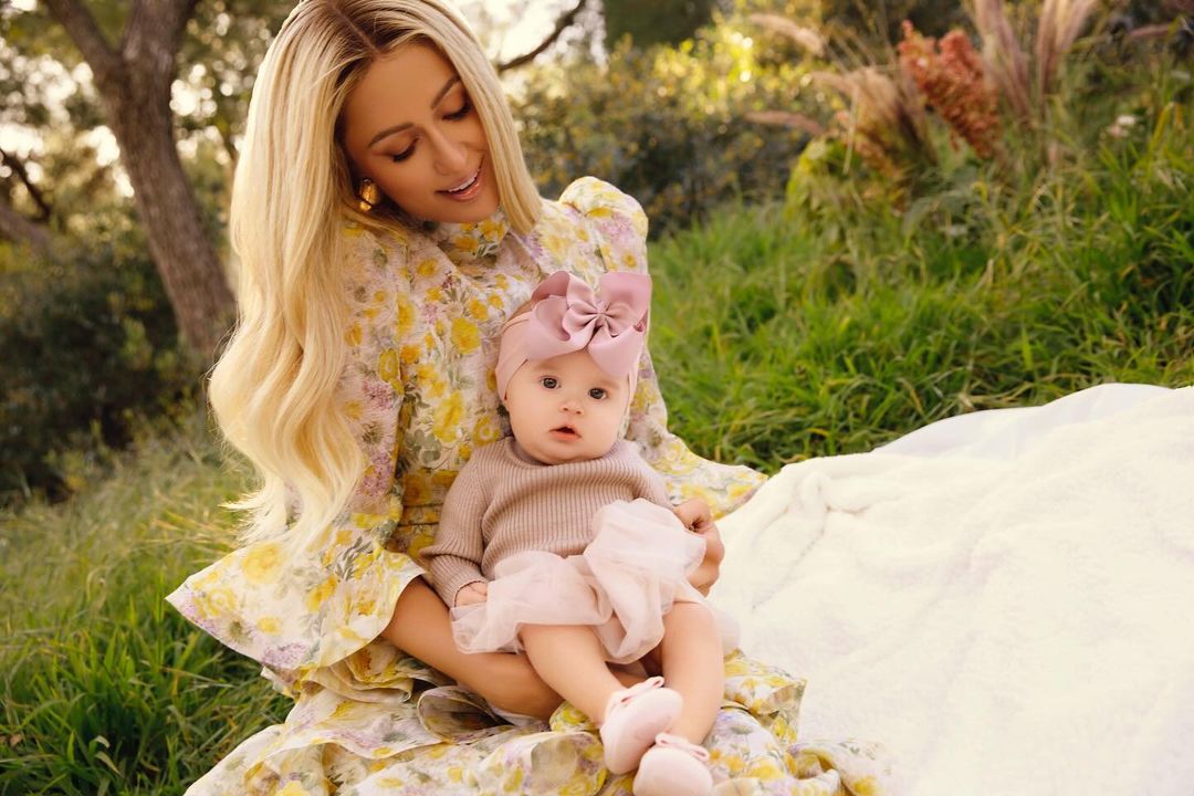 Paris Hilton poses outdoors with her baby girl, London, wearing a floral dress, with soft natural light and greenery in the background.