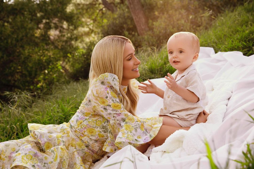 Woman and child sit together on a white blanket in a grassy field. The woman is wearing a floral dress and the child is wearing a beige outfit.