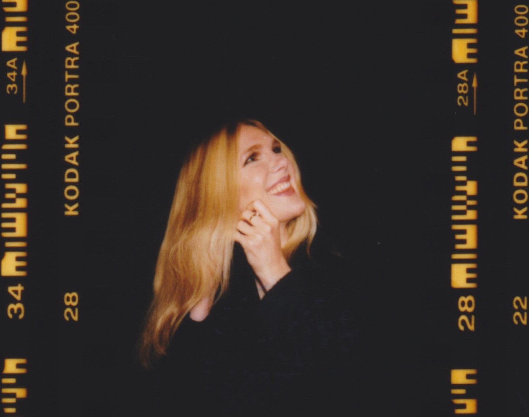 Portrait of a smiling woman with long blonde hair, fair skin, wearing a dark top, shot with Kodak Portra film; dark background.