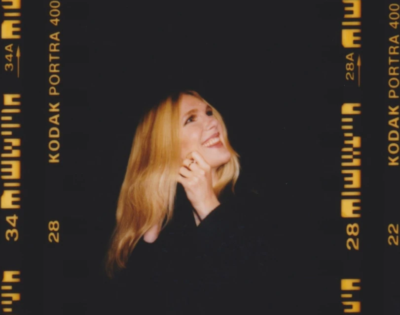 Portrait of a smiling woman with long blonde hair, fair skin, wearing a dark top, shot with Kodak Portra film; dark background.