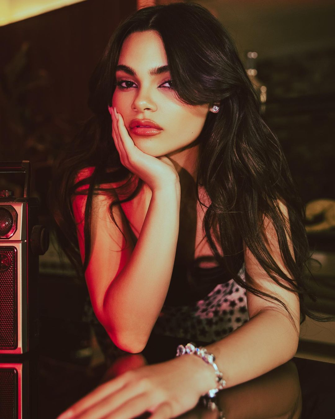 Brunette woman poses indoors, leaning on her hand with a vintage radio in the background, lit by soft, warm light.