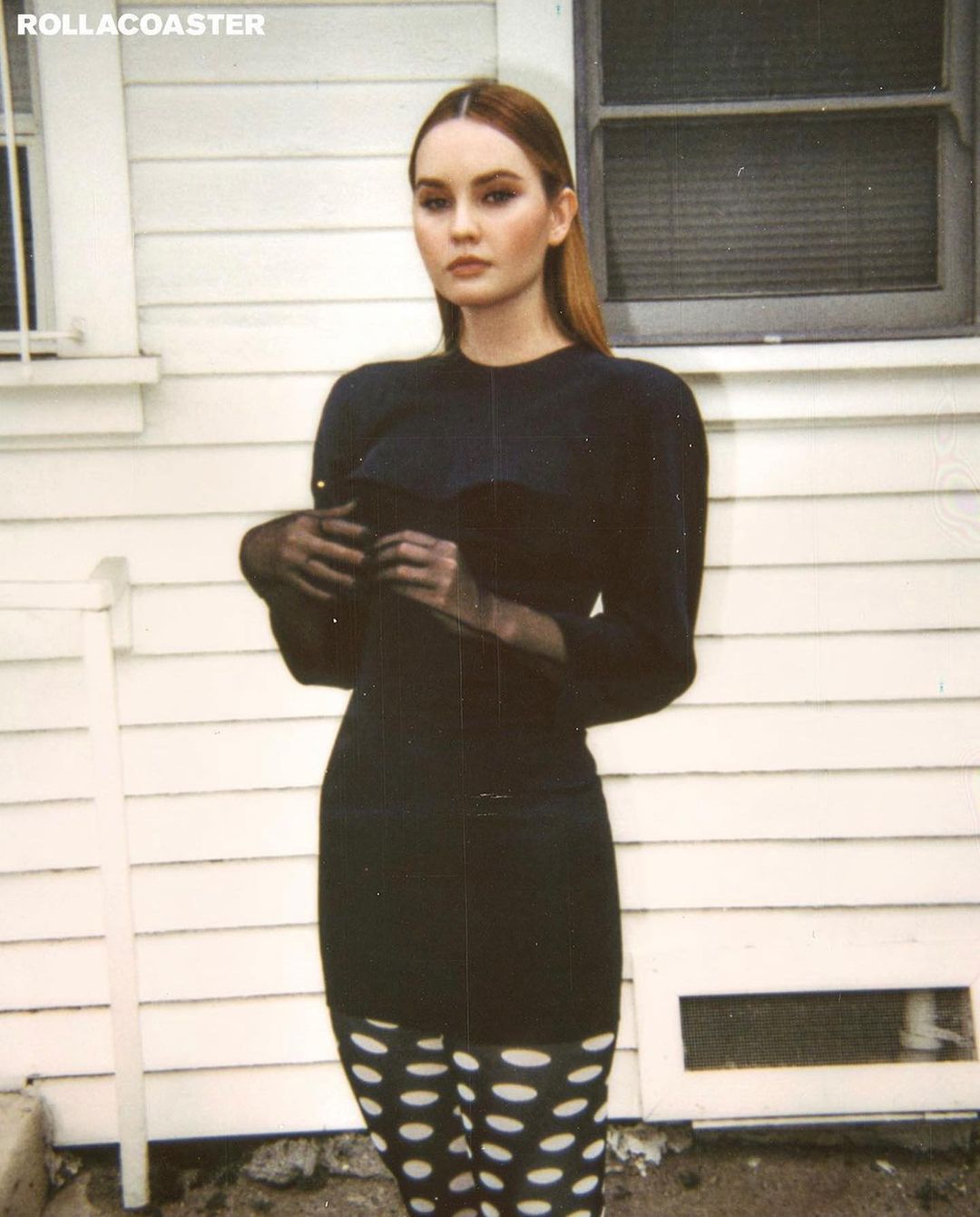 A young woman stands outside a white building wearing a black dress, sheer black gloves, and white tights with black polka dots.