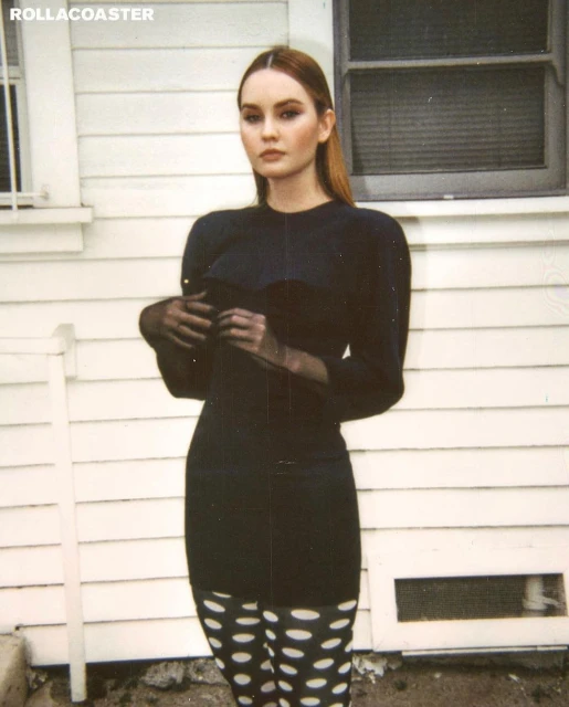 A young woman stands outside a white building wearing a black dress, sheer black gloves, and white tights with black polka dots.