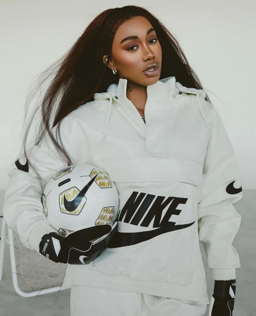 Woman in white Nike tracksuit holding soccer ball indoors. Female model with long dark hair in a studio-lit fashion pose.