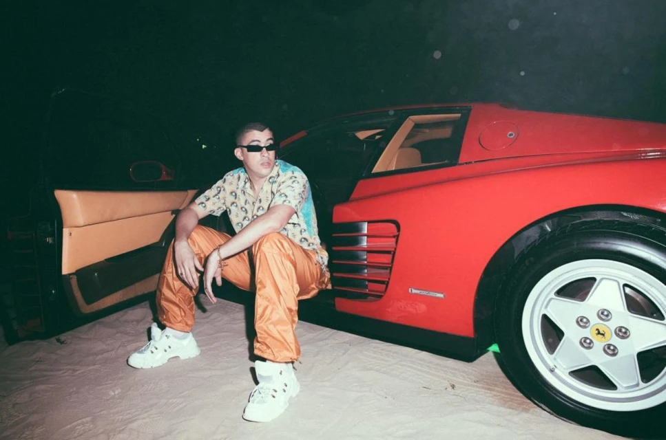 Bad Bunny poses beside a red Ferrari, wearing a patterned shirt, orange pants, and white sneakers in an outdoor nighttime setting.