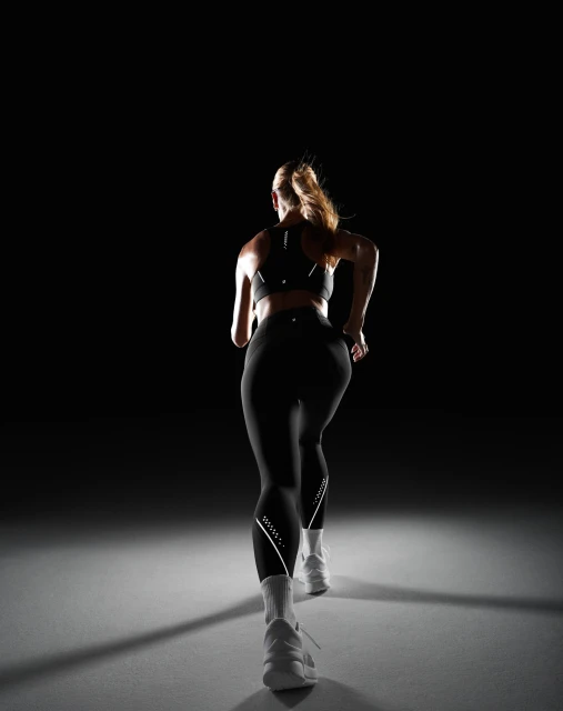 Rear view of a woman in workout attire running toward the viewer in a dimly lit photo studio with a dark background and grey flooring.