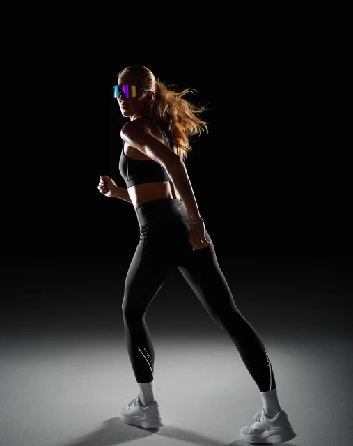 Athletic woman in black sportswear and futuristic visor-like sunglasses running in a dimly lit studio against a dark backdrop.