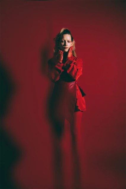 Woman in red outfit poses in front of a matching red backdrop with dramatic lighting and a vertical composition in a studio setting.