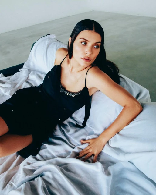 Woman with dark hair in black dress poses on bed with white sheets, glancing to the side in a fashion-style portrait.