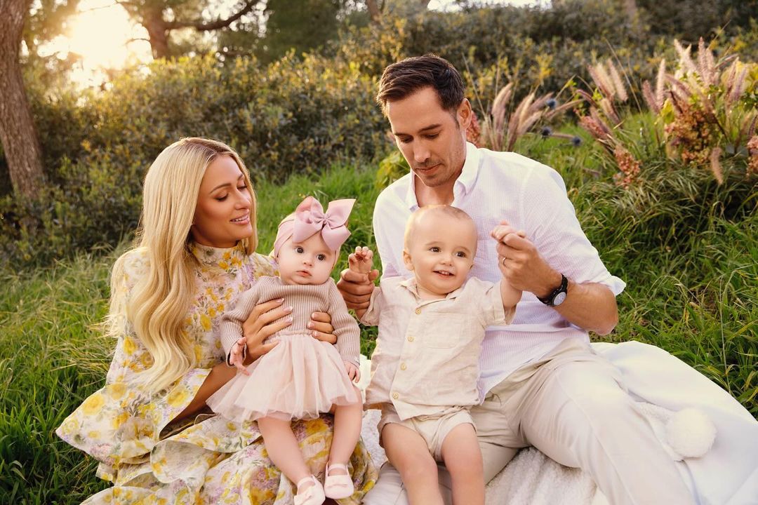 Paris Hilton with husband and two babies outdoors, sitting on blanket surrounded by green grass and trees in the background.