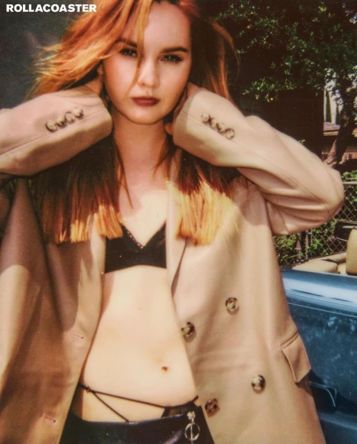 Attractive redhead woman poses in an open beige overcoat with a black bra and skirt, outdoors in daylight, shot at eye-level angle.