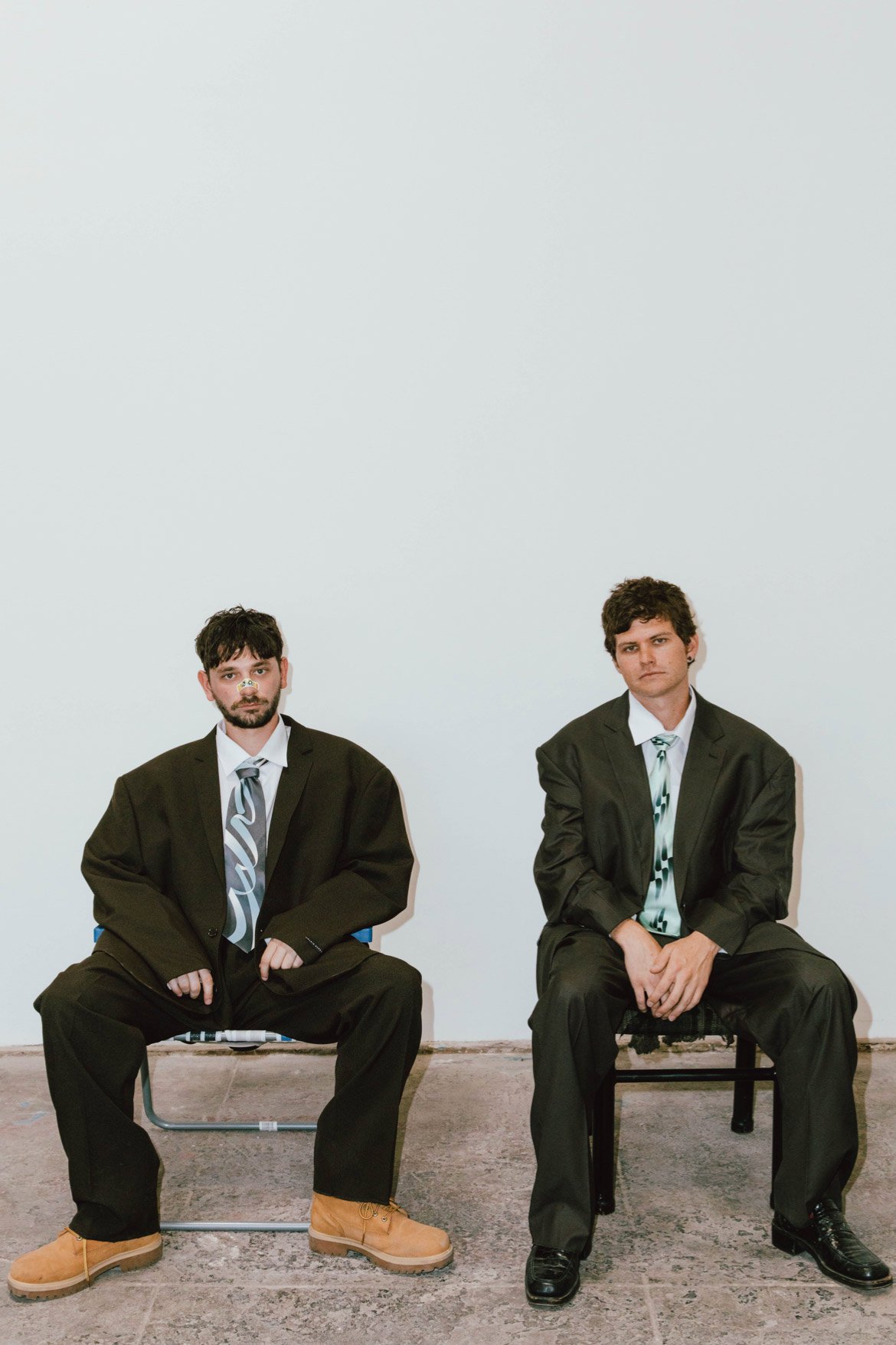 Two adult men sit on chairs in a bright room wearing oversized suits and ties. The men are looking at the camera.