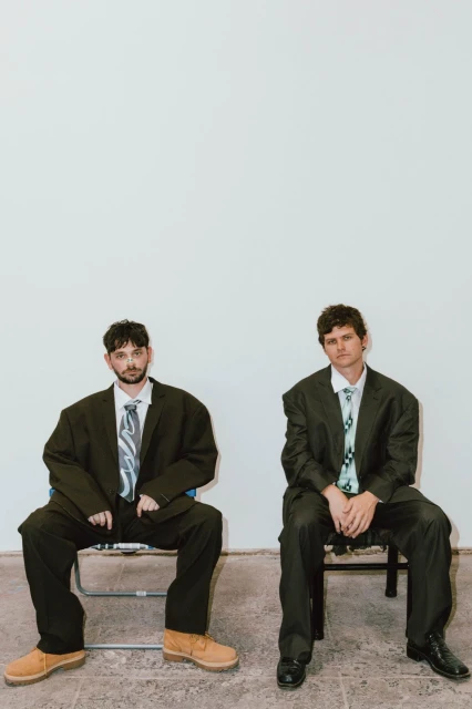 Two adult men sit on chairs in a bright room wearing oversized suits and ties. The men are looking at the camera.