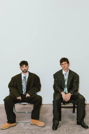 Two adult men sit on chairs in a bright room wearing oversized suits and ties. The men are looking at the camera.