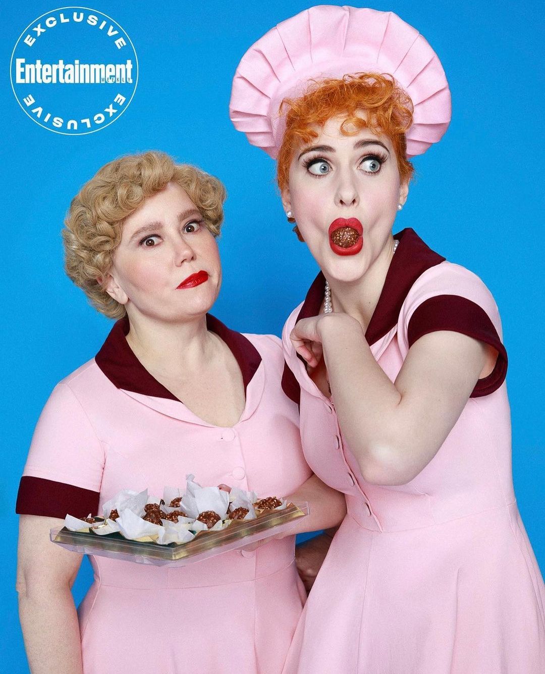 Two women dressed in pink dresses, styled as Lucille Ball characters, pose against a bright blue backdrop for an Entertainment Exclusive photo.