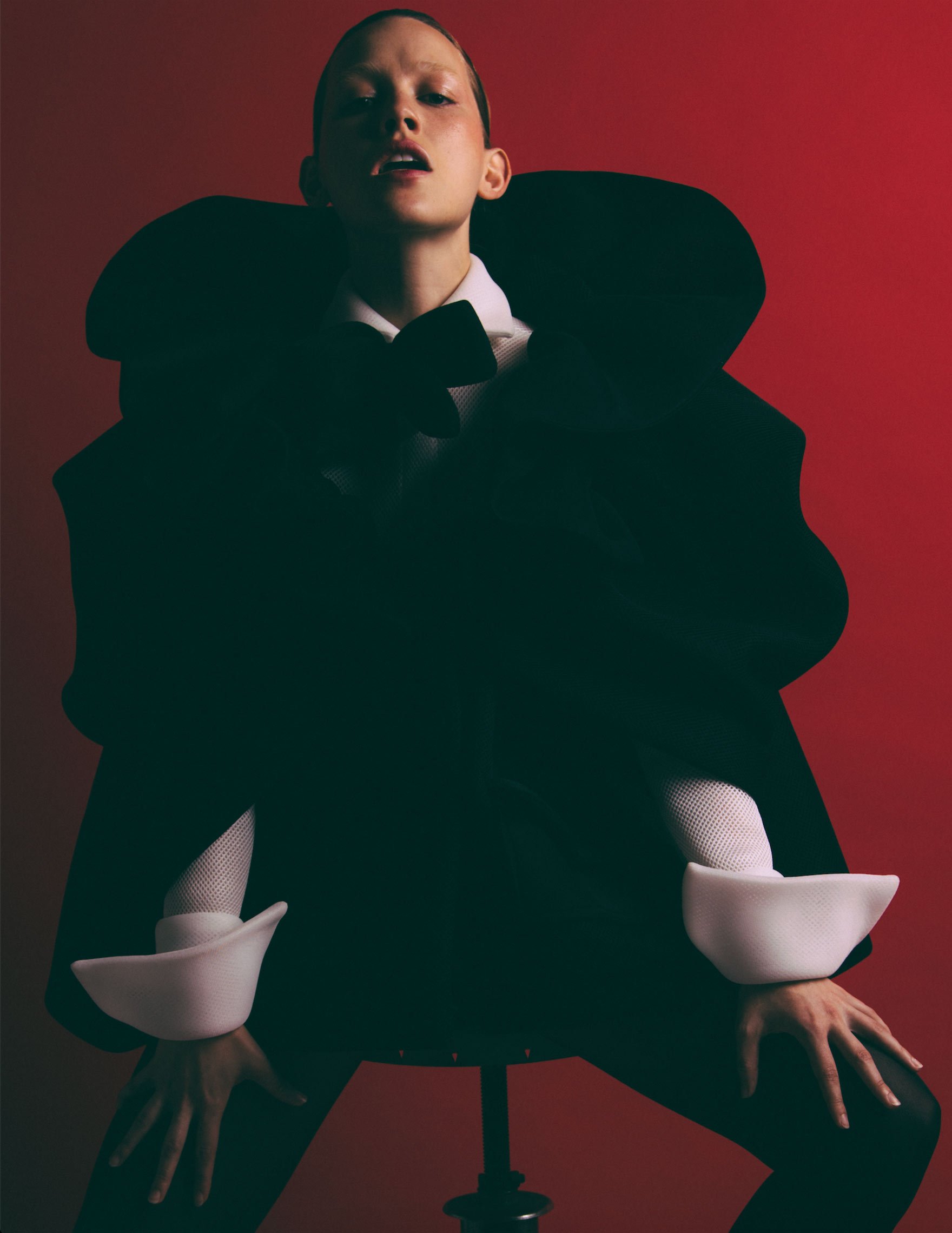 Woman poses in a dramatic black coat, bow tie, and white cuffs against a red background, seated on a stool in a studio.