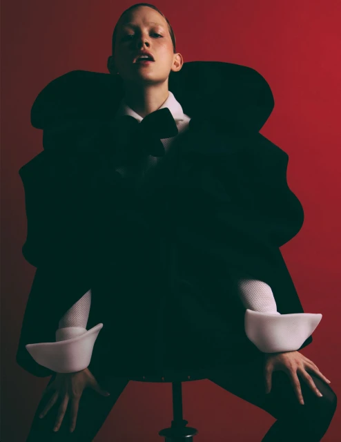 Woman poses in a dramatic black coat, bow tie, and white cuffs against a red background, seated on a stool in a studio.