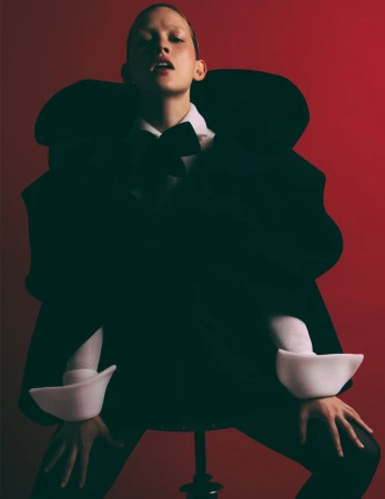 Woman poses in a dramatic black coat, bow tie, and white cuffs against a red background, seated on a stool in a studio.