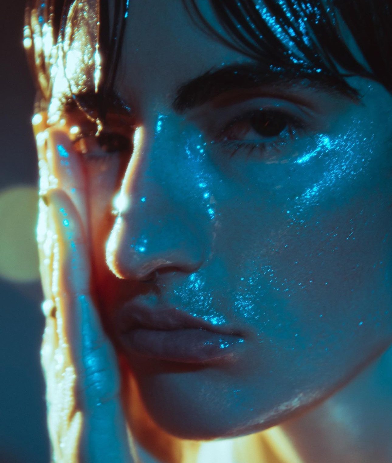 Close-up portrait of a woman with glittering blue makeup on her face and hair, softly lit and emphasizing textures.