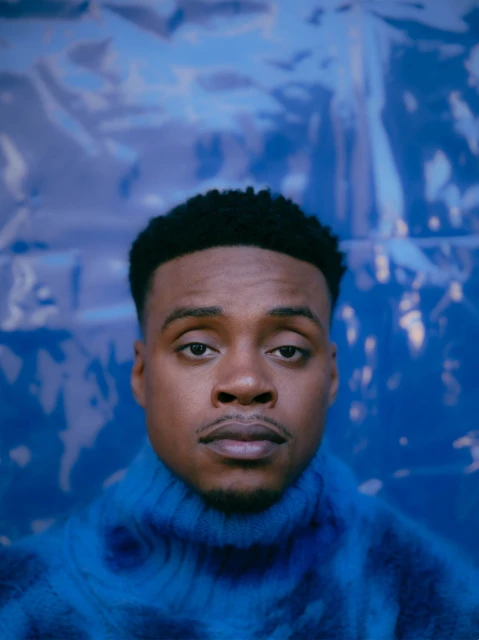 Portrait of a young African-American man in a blue patterned turtleneck sweater against a crinkled blue foil background in soft lighting.