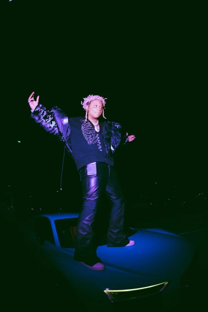 A young adult stands on a car under a dark night sky, wearing a black sweater, black leather pants, and colorful dreadlocks.