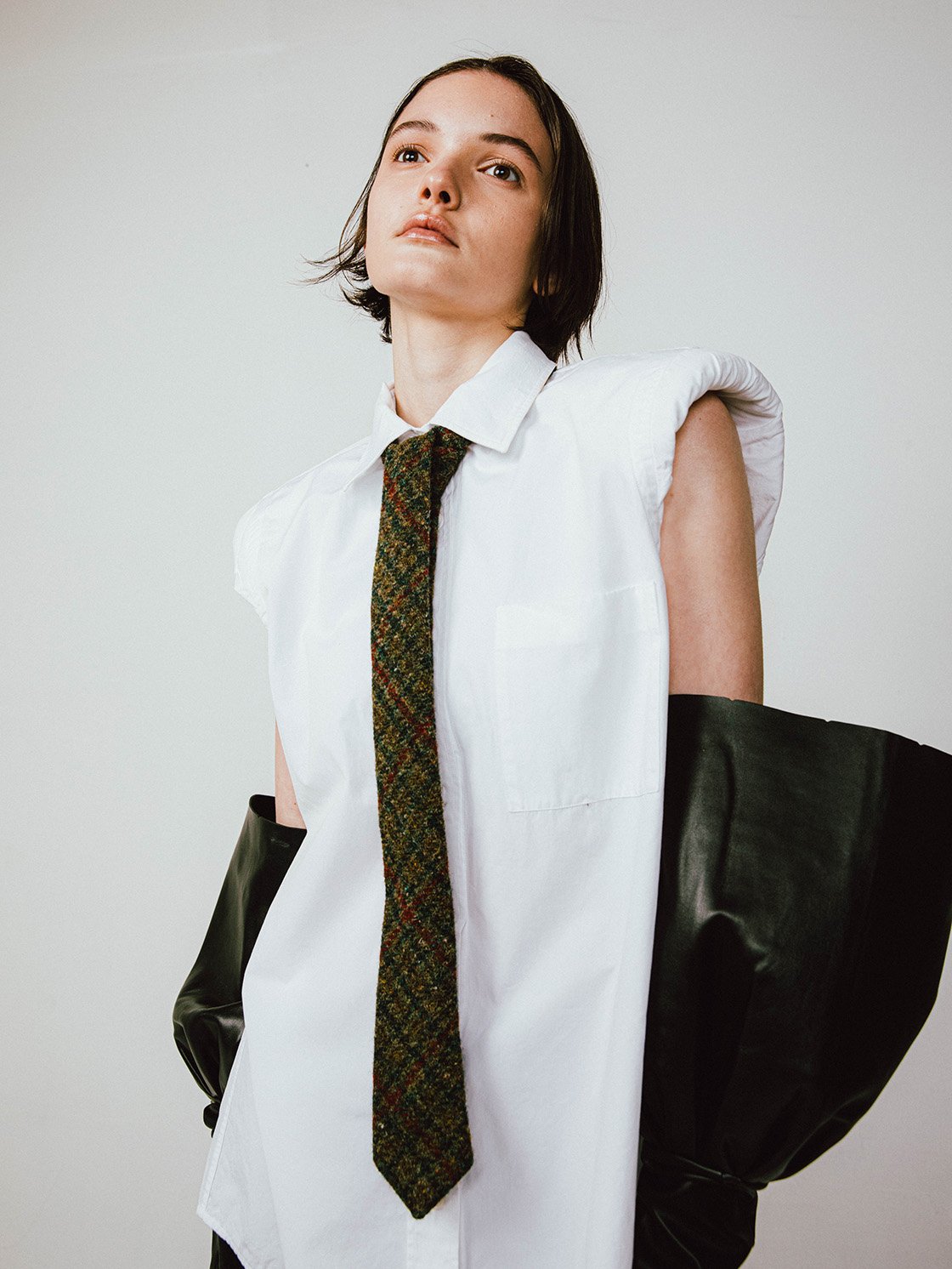 A woman poses in a white, oversized shirt and knit tie, paired with black elbow gloves against a neutral studio backdrop in portrait orientation.