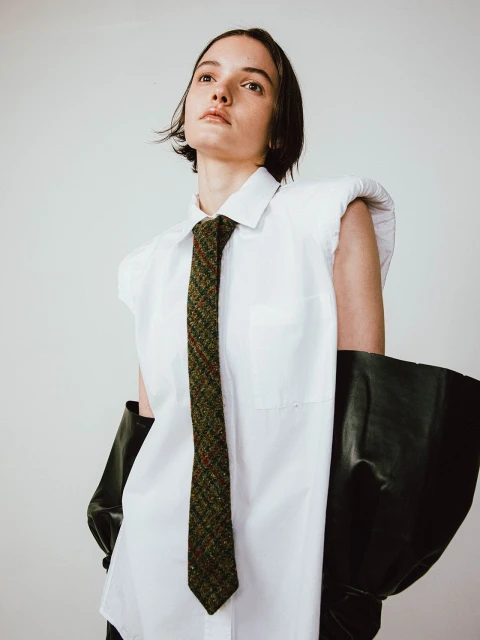 A woman poses in a white, oversized shirt and knit tie, paired with black elbow gloves against a neutral studio backdrop in portrait orientation.