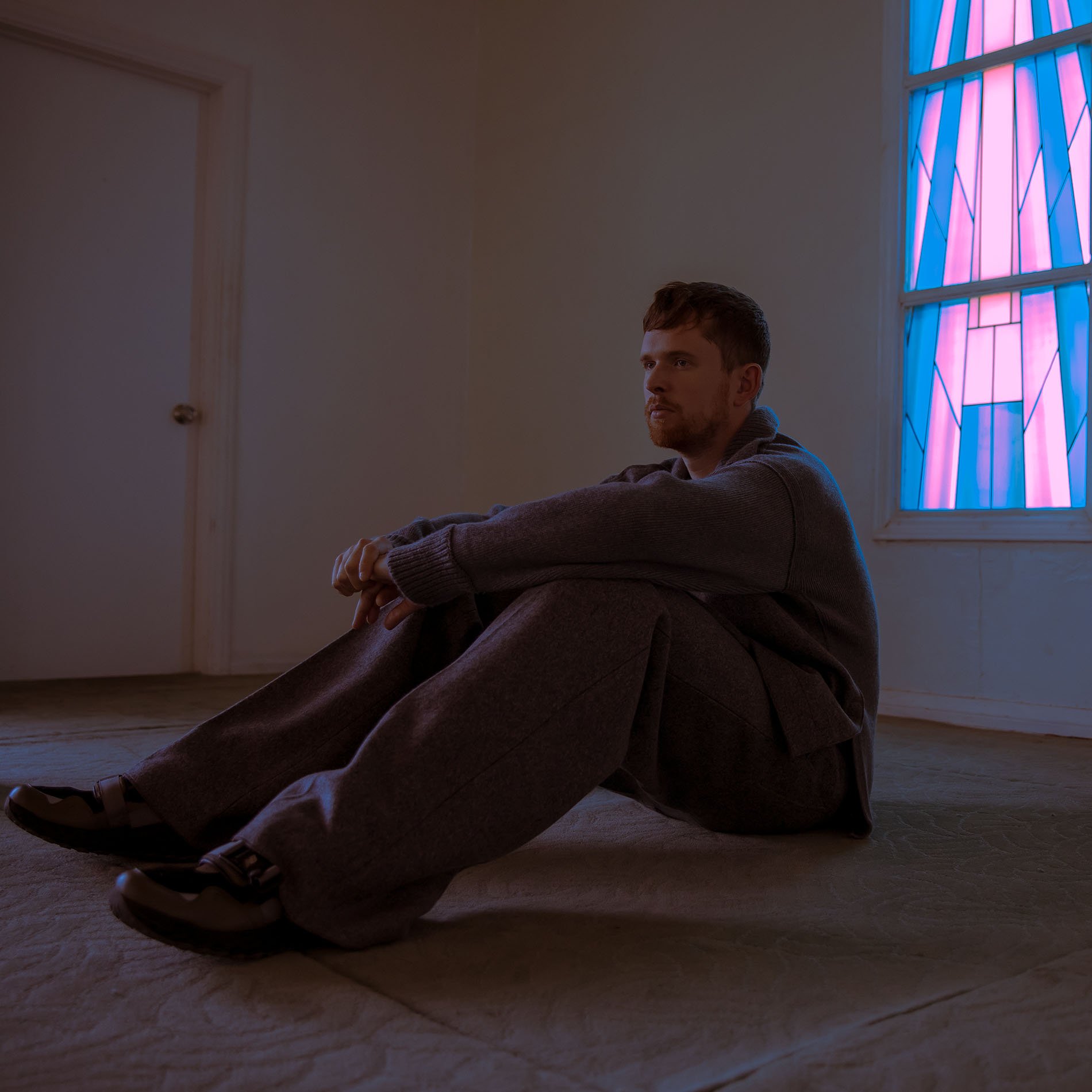 Adult male in grey loungewear sitting on the floor in front of a blue and pink stained-glass window and a white door in a simple room.