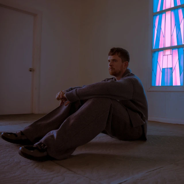 Adult male in grey loungewear sitting on the floor in front of a blue and pink stained-glass window and a white door in a simple room.
