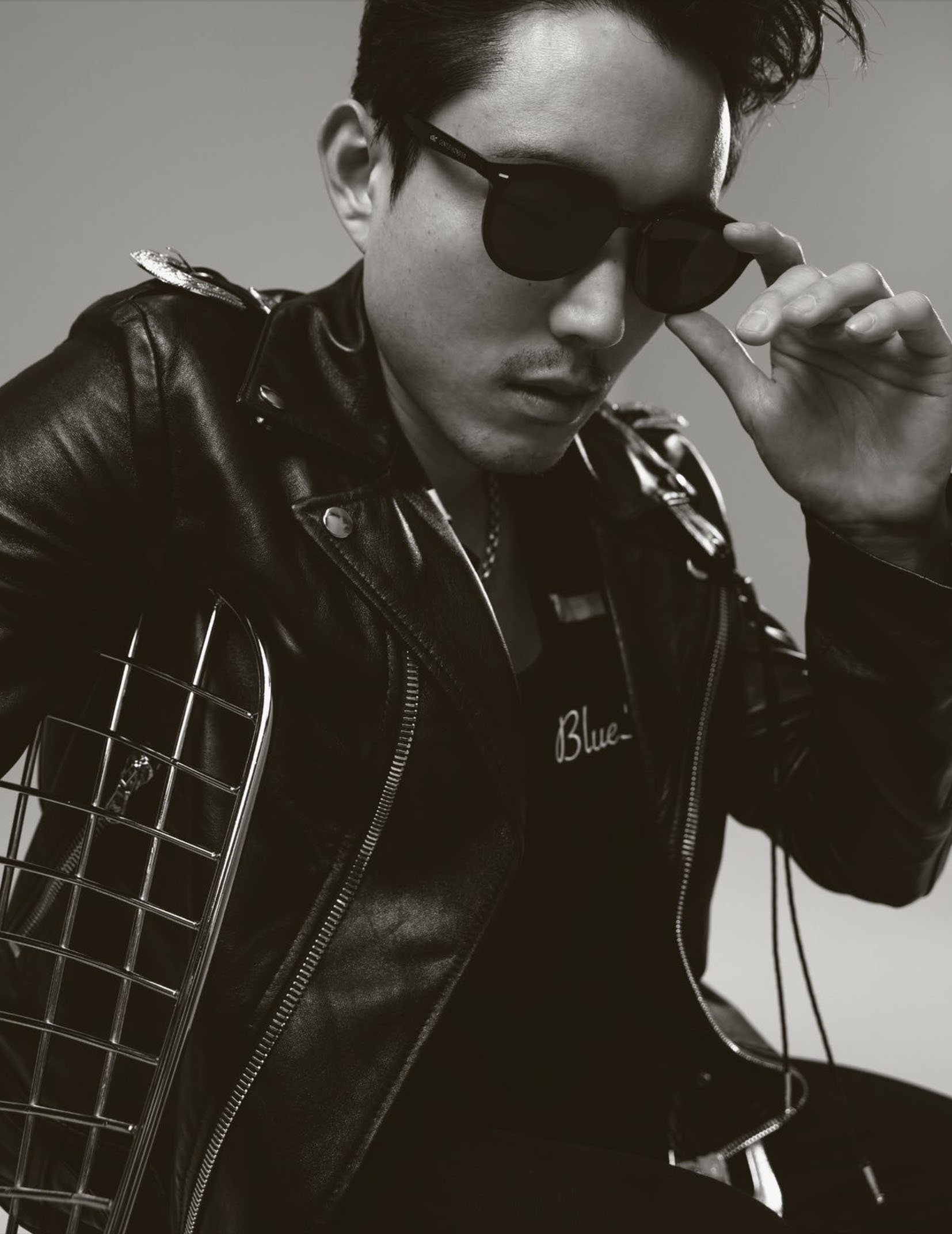 Monochrome portrait of an adult male wearing sunglasses and a leather jacket, sitting in a metal chair against a plain background.