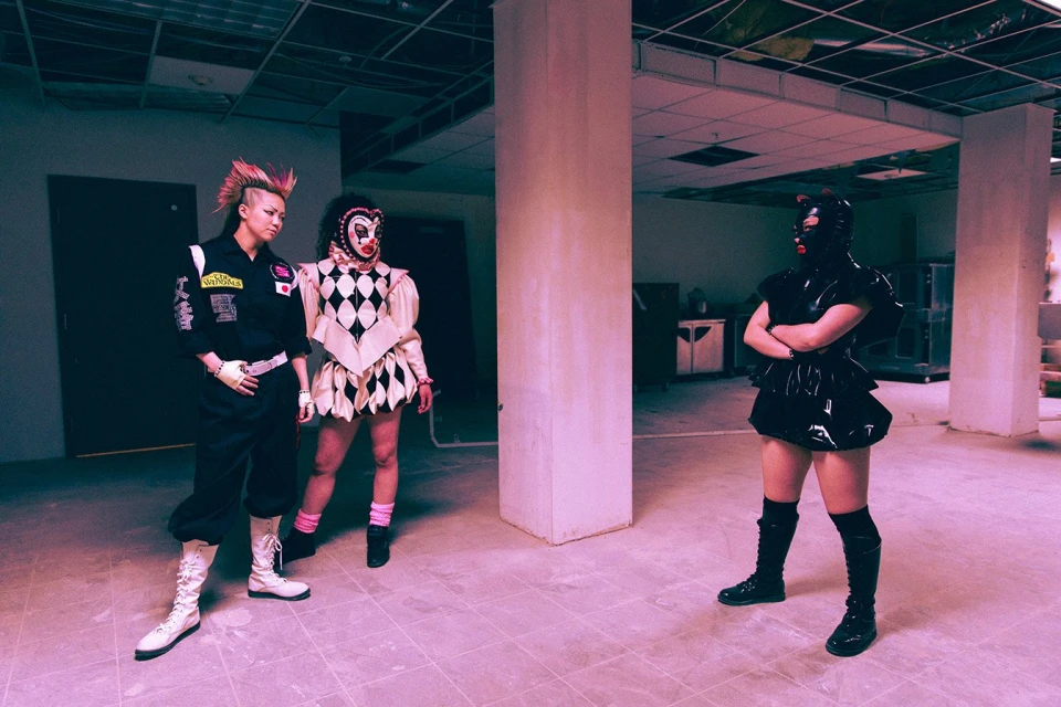 Three figures in a sparsely decorated room. One is wearing a black catsuit, another is in a clown costume, and a third in a black jumpsuit.