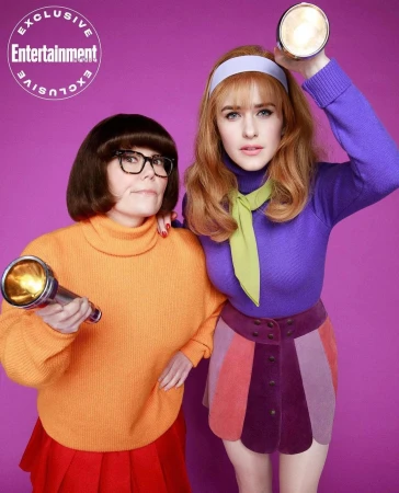Two women in costumes holding flashlights pose against a purple backdrop; one with auburn hair, the other with dark bob. Exclusive Entertainment logo visible.