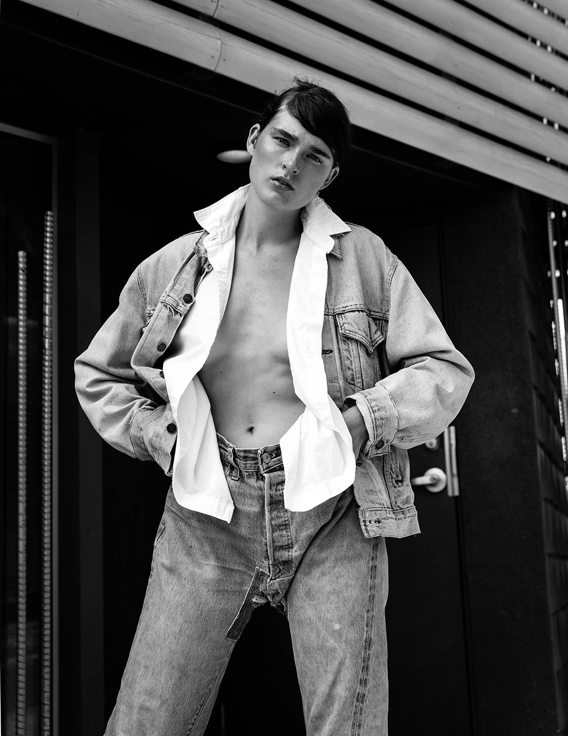 Black and white portrait of a androgynous person wearing a denim jacket and jeans standing in front of a dark building exterior.