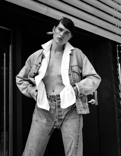 Black and white portrait of a androgynous person wearing a denim jacket and jeans standing in front of a dark building exterior.