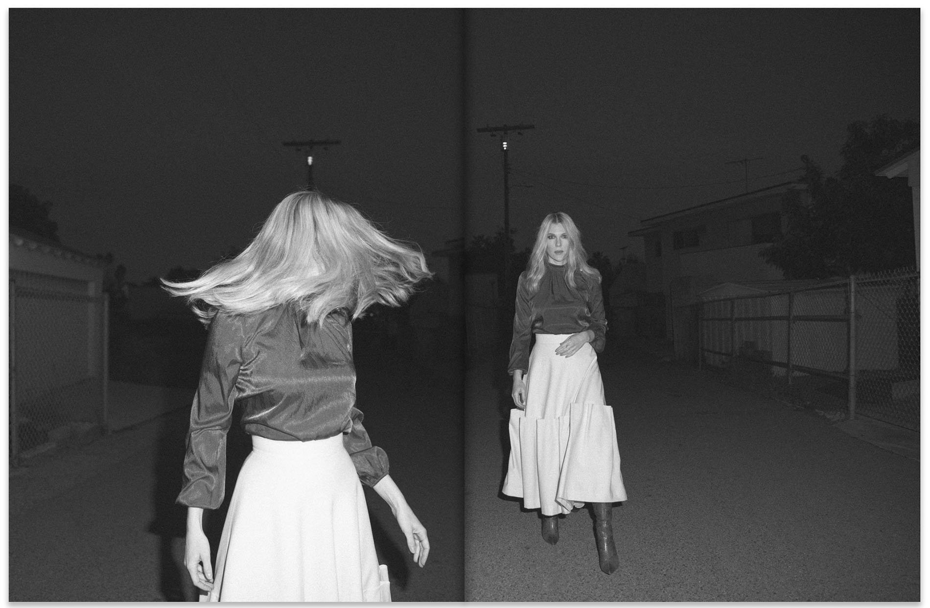 Two women wearing skirts and blouses stand on a road at night. One woman poses while the other whips her hair.