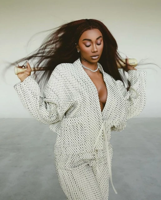 Woman in a patterned jacket and pants with long hair, wearing a chain necklace, holding a gold tube, posing in a neutral setting.