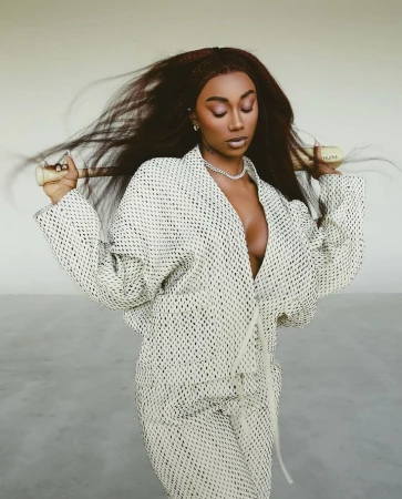 Woman in a patterned jacket and pants with long hair, wearing a chain necklace, holding a gold tube, posing in a neutral setting.
