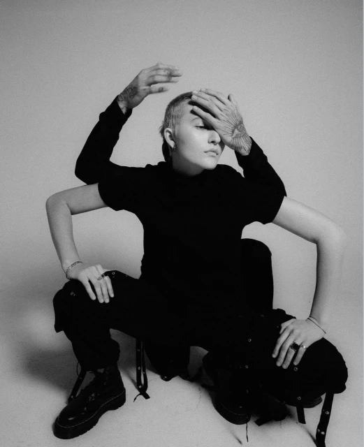 Black and white studio portrait of a young adult with short hair and tattoos posing with hands near face in dark monochrome clothing.