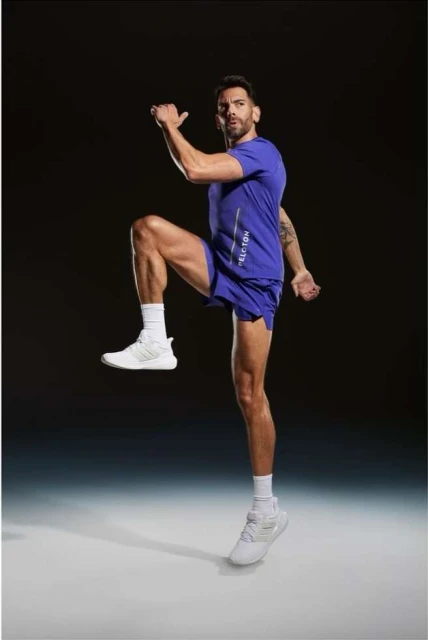 Athletic man in blue activewear mid-air, performing a high knee exercise with focused expression against a dark backdrop in studio lighting.