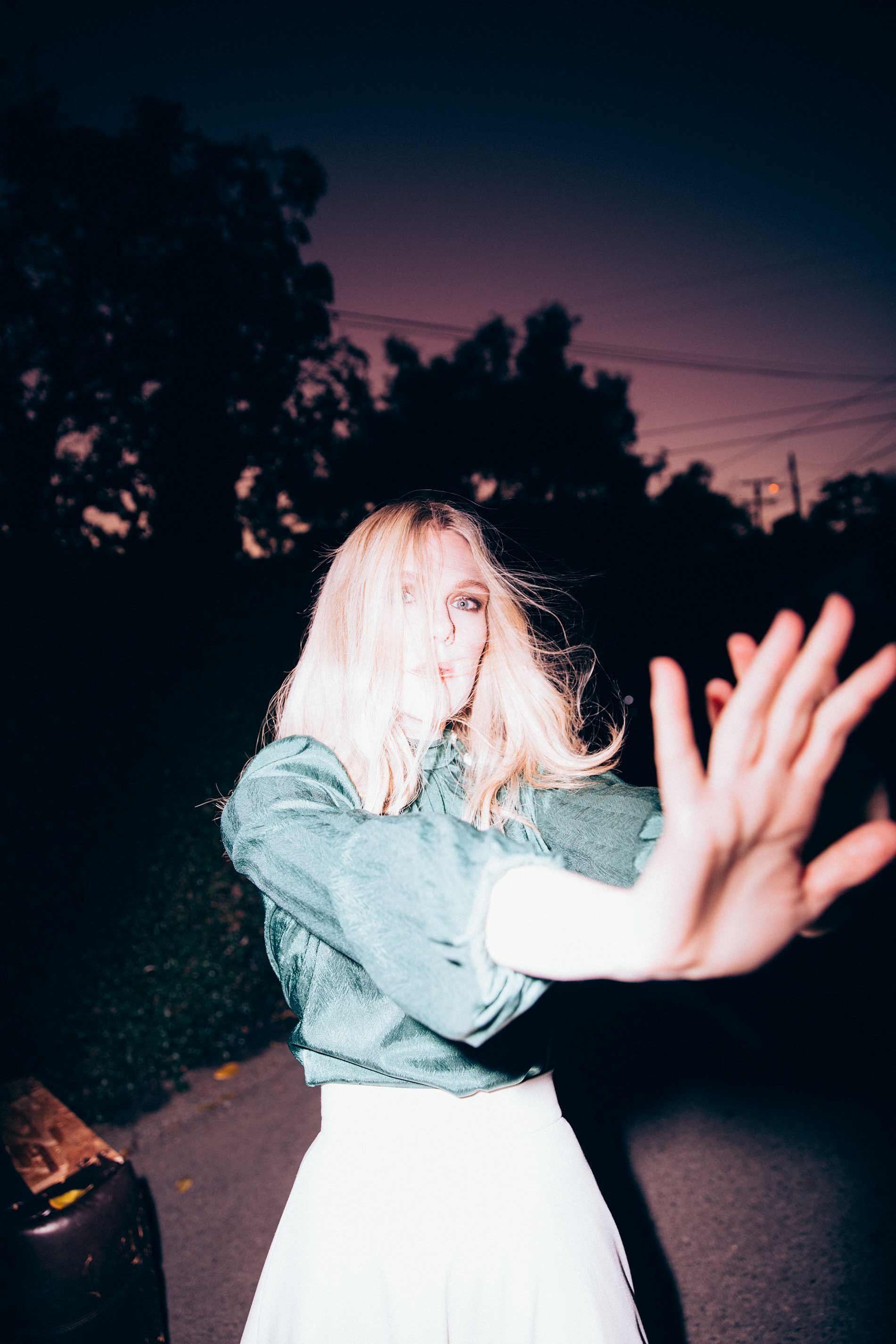Woman with blonde hair poses in front of dark foliage and street at dusk wearing an aqua top and a white skirt with outstretched arms.