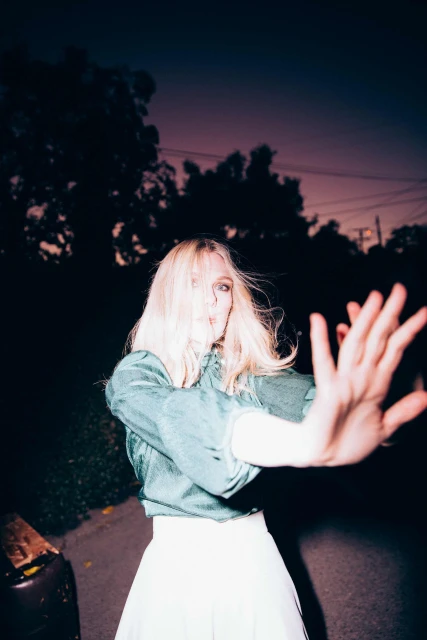 Woman with blonde hair poses in front of dark foliage and street at dusk wearing an aqua top and a white skirt with outstretched arms.