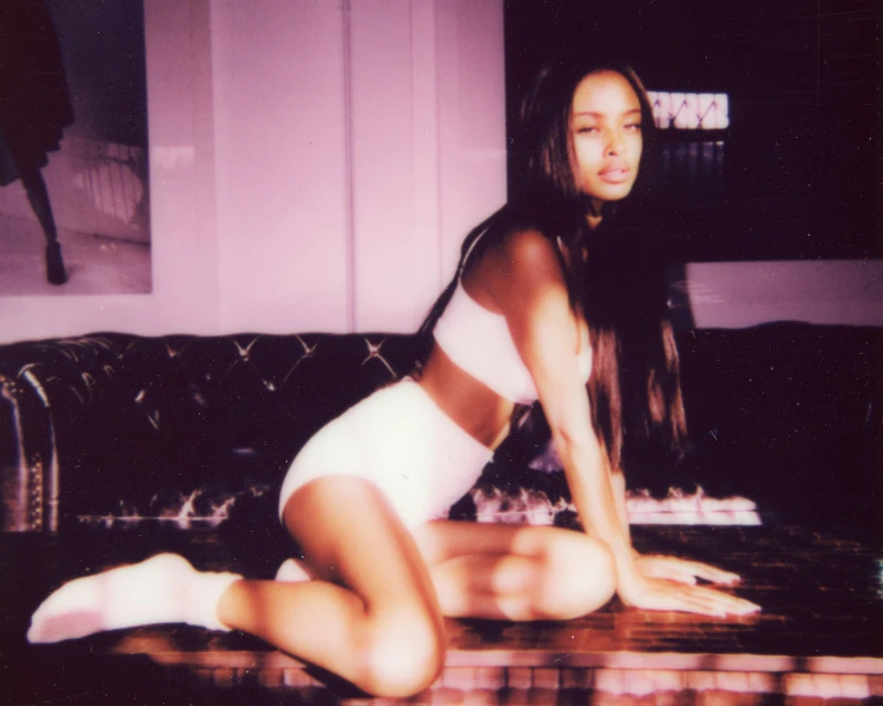 Indoor portrait of a Black woman with long hair, wearing a bra top and shorts, kneeling on a dark, tufted surface in a studio setting.